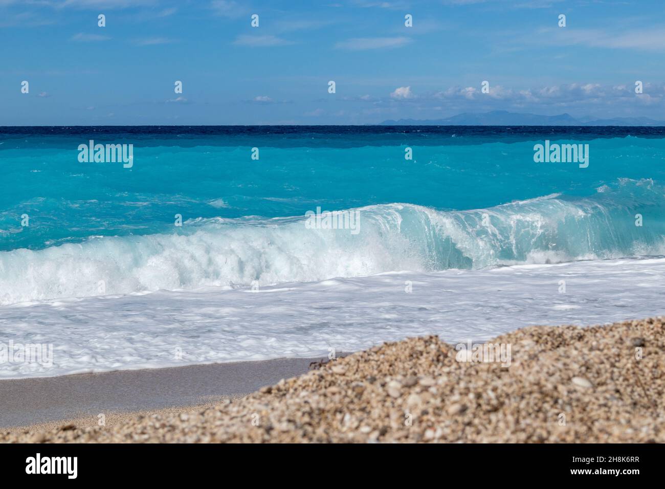 Azure Vibrant Breaker onde colpire ghiaia riva. Costa soleggiata dell'isola greca con cielo blu. Spiaggia di sabbia in Grecia. Viaggio estivo nella natura verso il Mar Ionio Foto Stock