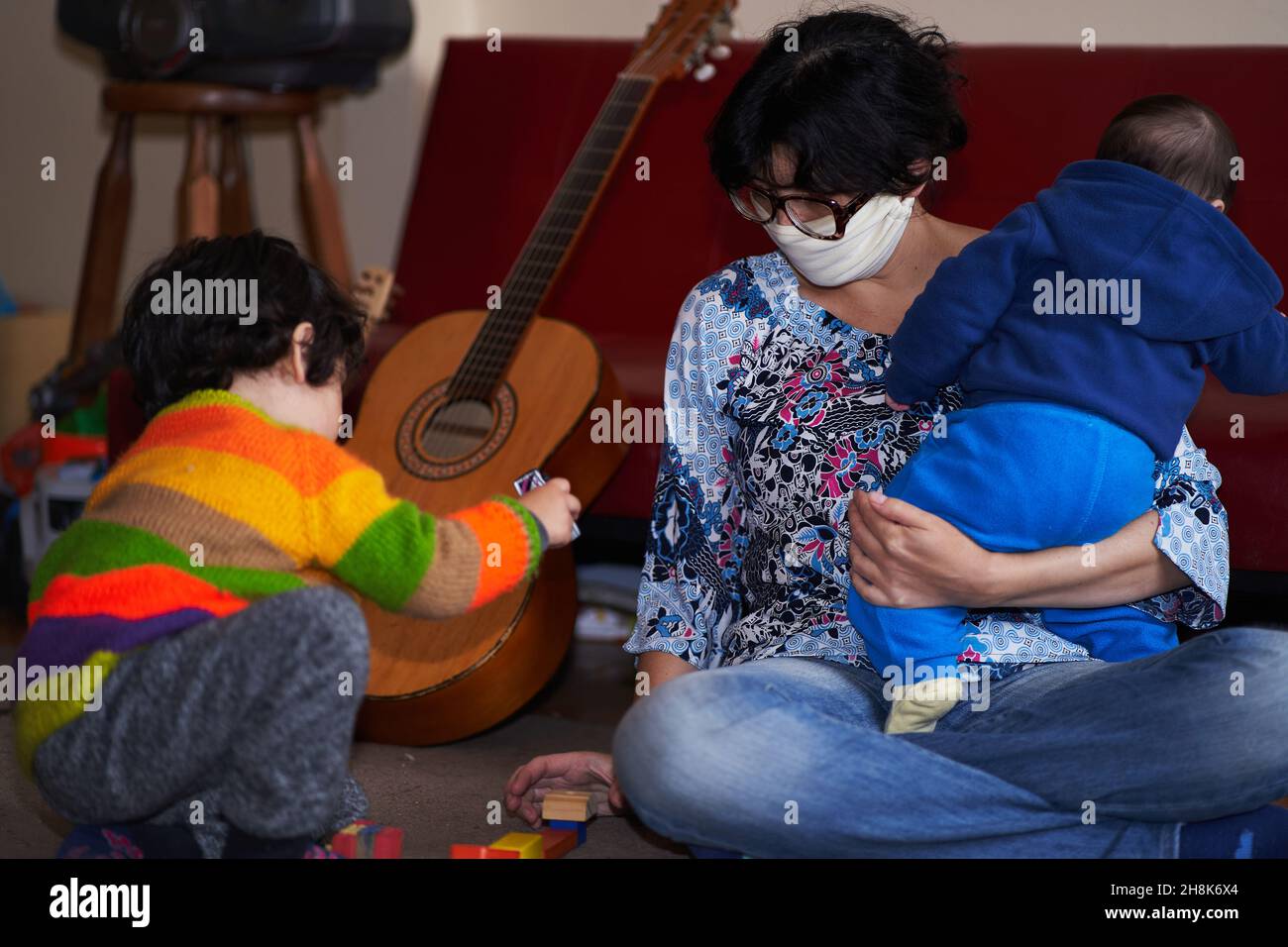 donna con maschera passa la chitarra al bambino mentre tiene il bambino in braccio. giochi pandemici a casa. covid-19 coronavirus Foto Stock