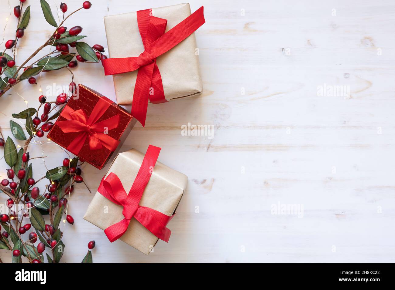 Tre regali di Natale con fiocchi a nastro rossi su sfondo di legno bianco con bordo di bacche di agrifoglio rosse e mini luci bianche in posa piatta, spazio copia Foto Stock