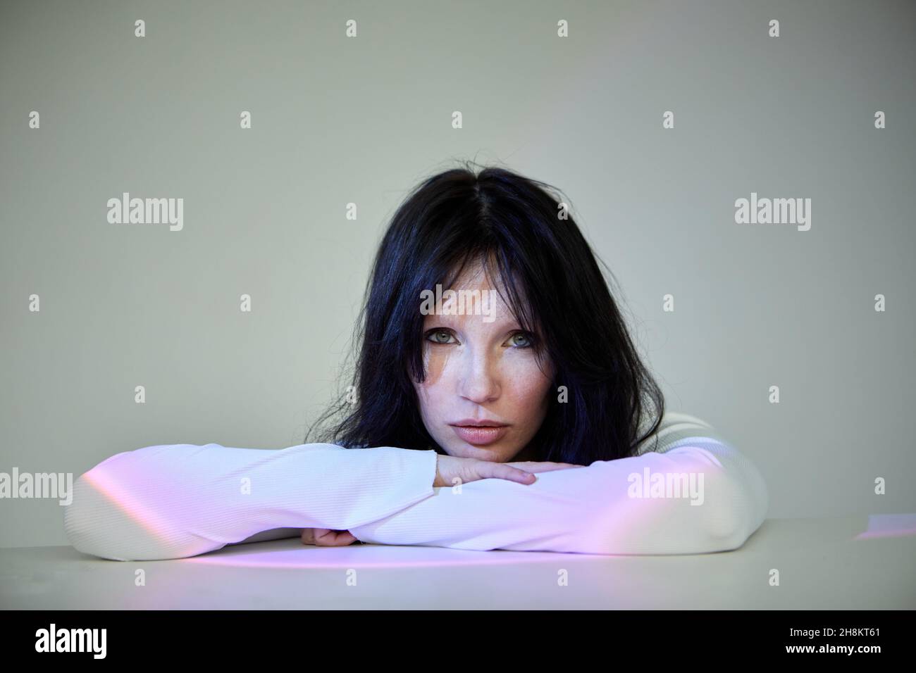 Donna seria ripiegando le mani sul tavolo e guardando la telecamera in raggi di illuminazione su sfondo grigio Foto Stock Donna seria ripiegando le mani sul tavolo e guardando la telecamera in raggi di illuminazione su sfondo grigio Foto Stock
