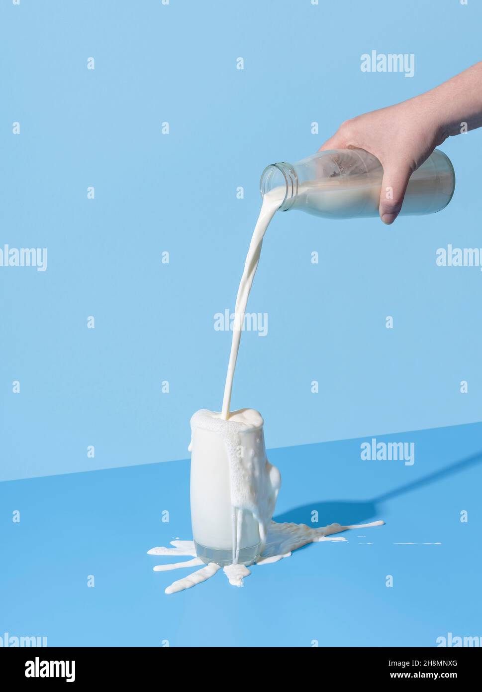 Donna a mano versando il latte da una bottiglia nel bicchiere, minimalista su sfondo blu. Eccesso di latte sulla tavola di colore blu. Foto Stock