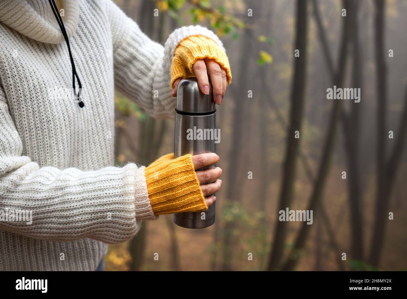 Thermos di apertura dell'escursionista con bevanda calda nella foresta. Donna che indossa maglione a maglia e guanti in lana senza dita. Rinfresco durante l'escursione all'aperto Foto Stock