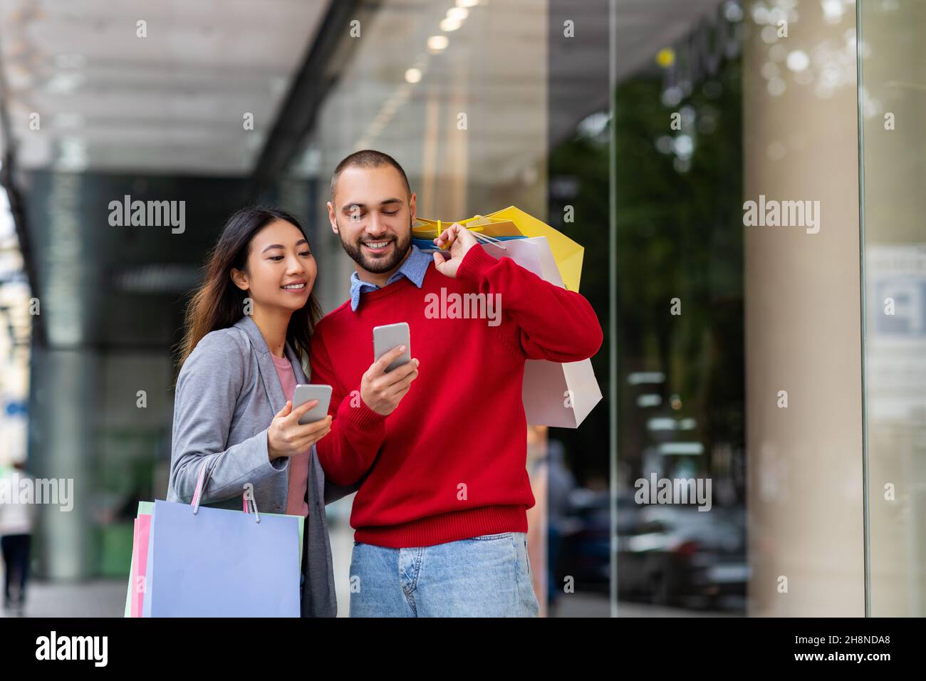 Giovane coppia interrazziale con smartphone in piedi su strada con borse shopping, acquistare merci online vicino al grande centro commerciale della città Foto Stock