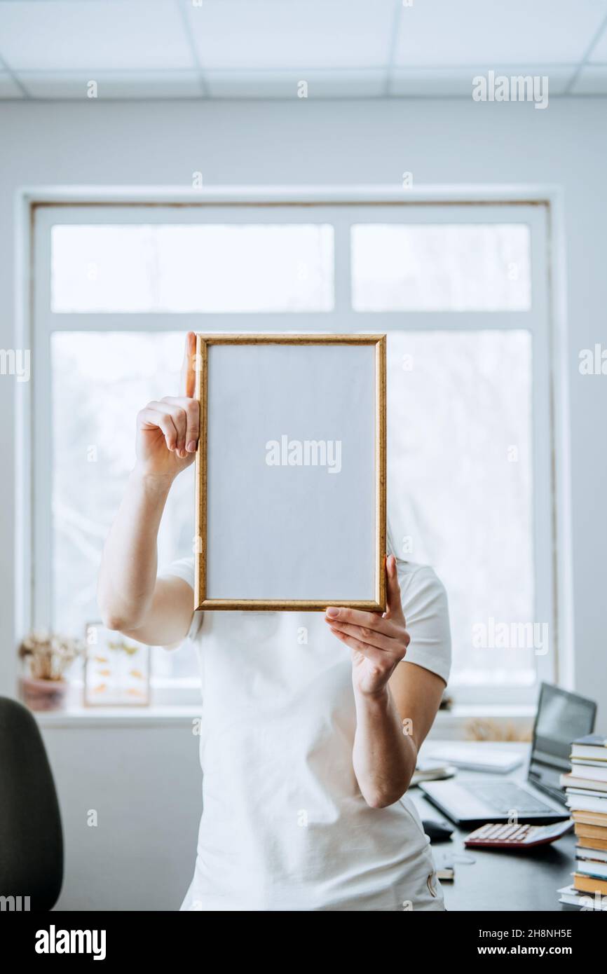 Cornice, poster, diploma, mockup certificato in mani femminili. Donna senza volto che tiene vuota cornice fotografica di legno bianco sul tavolo con il laptop e. Foto Stock
