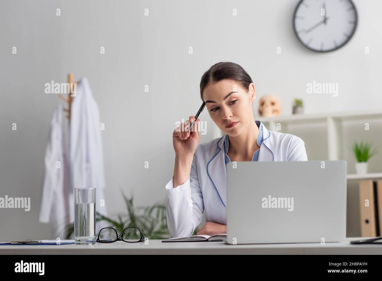 medico attento che guarda il computer portatile mentre si siede con la penna sul posto di lavoro Foto Stock