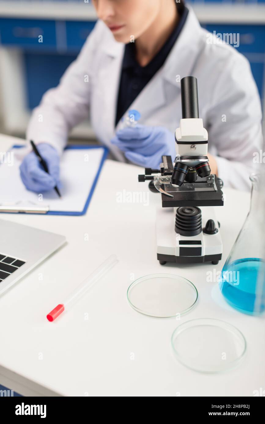 vista ritagliata della donna sfocata che scrive sulla clipboard vicino al microscopio e petri piatti sulla scrivania Foto Stock