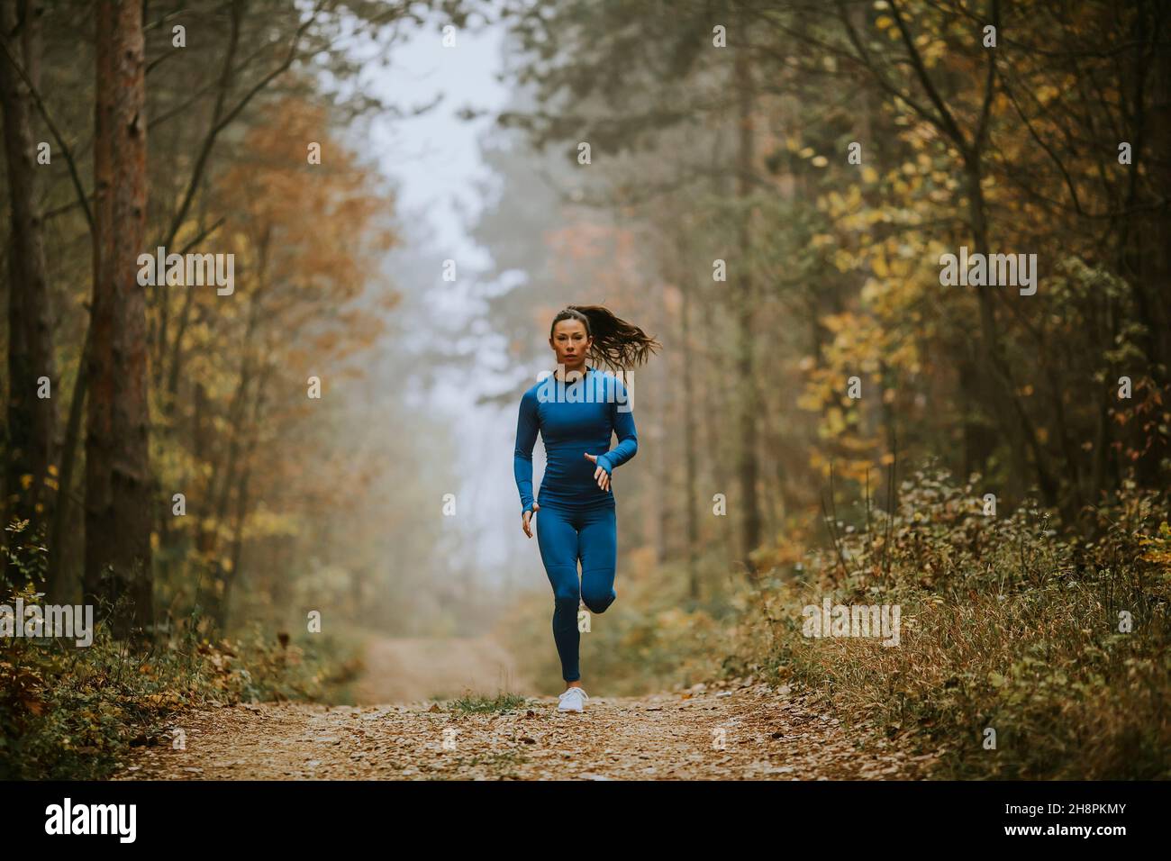 Giovane donna in tuta da pista blu che corre verso la macchina fotografica sul sentiero forestale in autunno Foto Stock