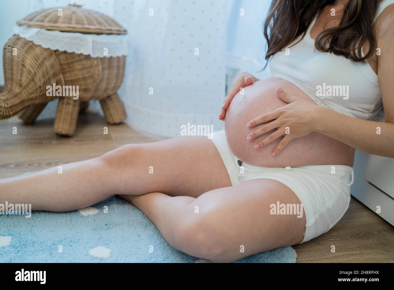 Donna incinta irriconoscibile carezzando il ventre mentre si siede sul pavimento Foto Stock