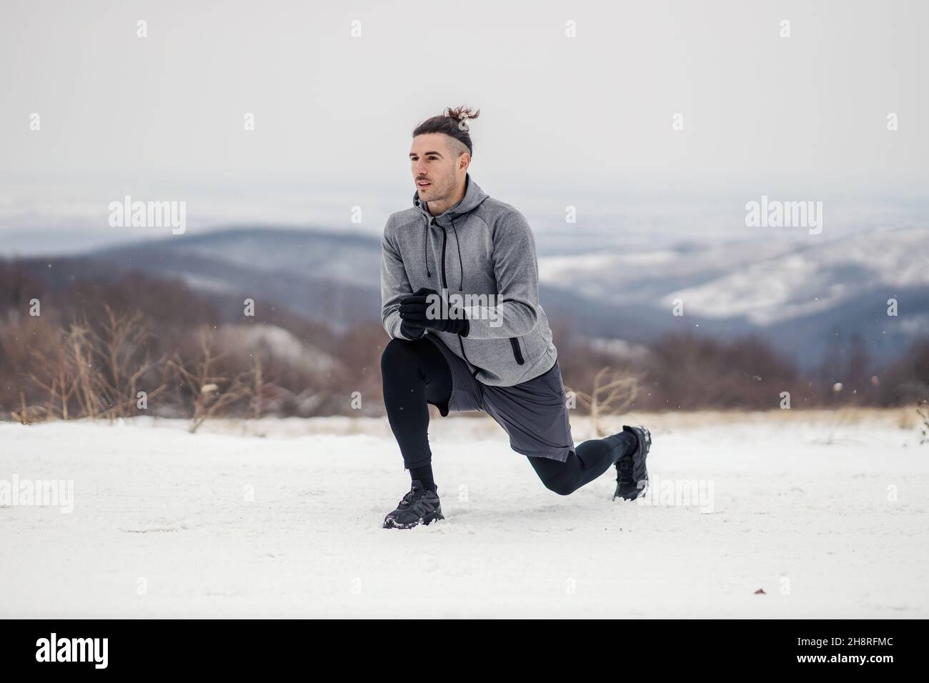Sportivo in forma inginocchiato e facendo esercizi di riscaldamento e stretching sulla neve in inverno. Fitness invernale, clima freddo, riscaldamento Foto Stock