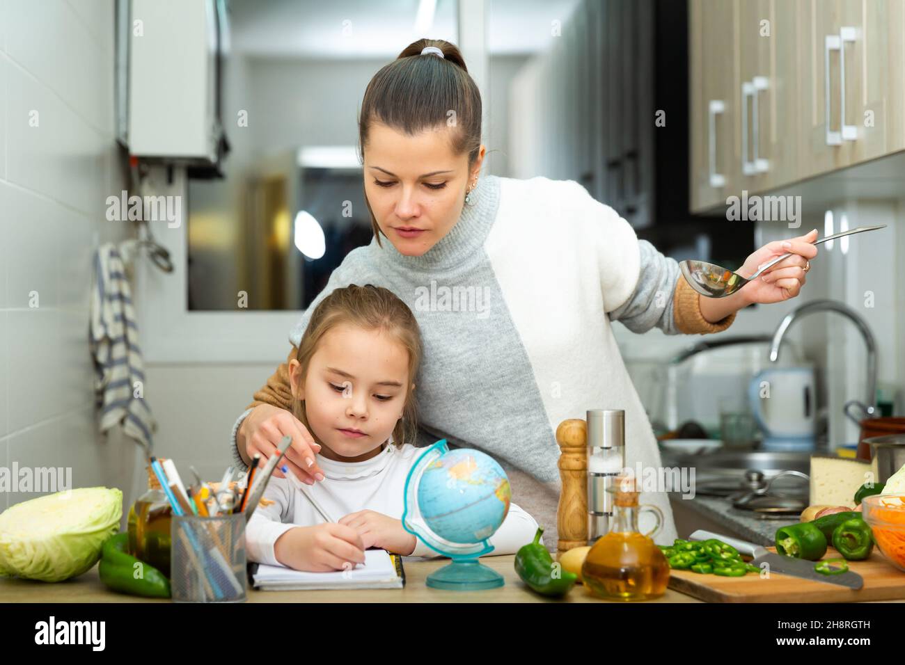 La madre che cucinava e la figlia piccola che faceva i compiti a scuola Foto Stock