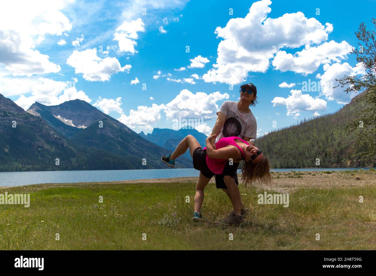 Divertente coppia gara mista posa sciocca in piedi sul prato di fronte alle montagne dal lago con cielo blu nuvole soffici Foto Stock