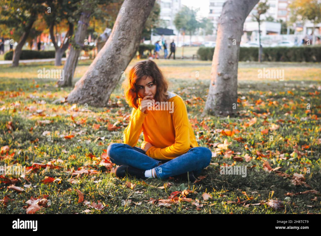 Giovane ragazza curly in maglione giallo e jeans seduti su erba caduta con foglie secche. Foto Stock