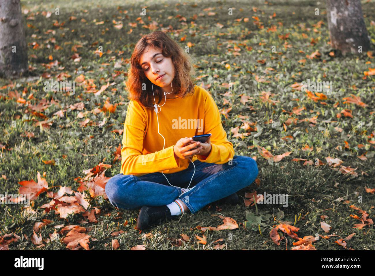 Giovane ragazza in maglione giallo che ascolta musica con auricolari seduti sull'erba d'autunno Foto Stock