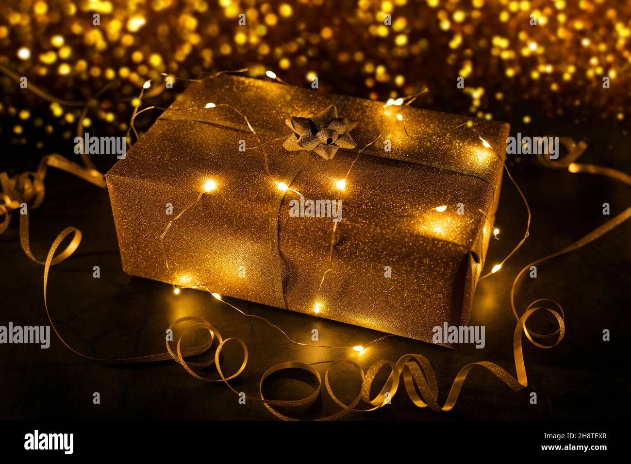 regalo d'oro avvolto in luci a corda su sfondo dorato bokeh Foto Stock