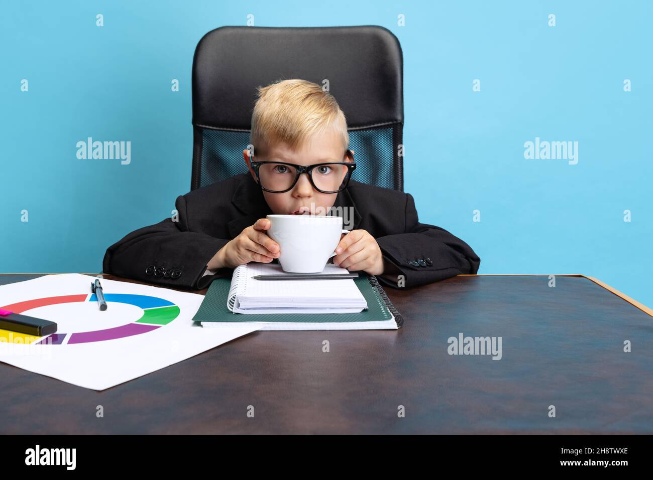 Ritratto di ragazzo piccolo, uomo d'affari bambino in giacca enorme vestito seduto al tavolo d'ufficio isolato su sfondo blu studio. Foto Stock