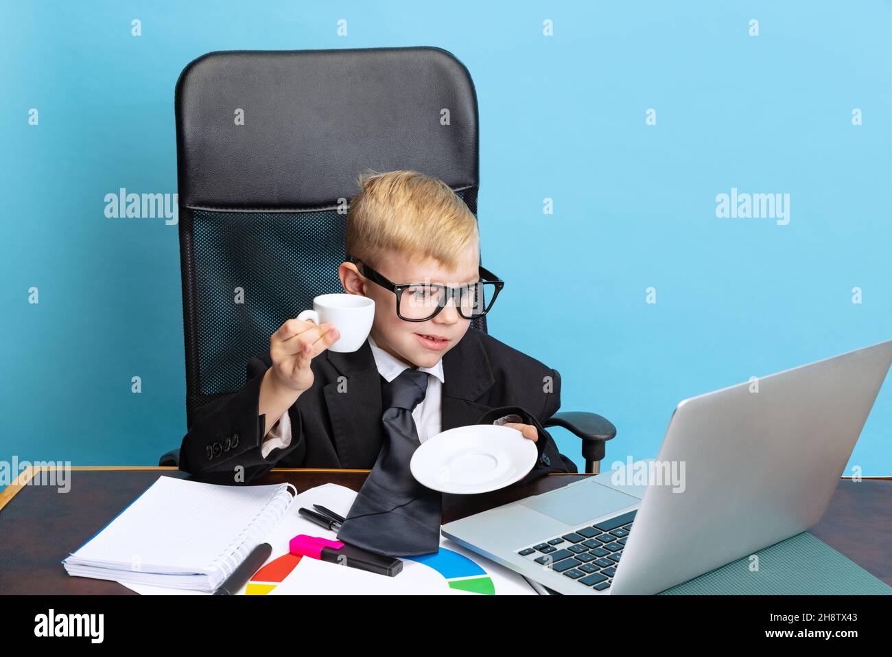 Ritratto di ragazzo piccolo, uomo d'affari bambino in giacca enorme vestito seduto al tavolo d'ufficio isolato su sfondo blu studio. Foto Stock
