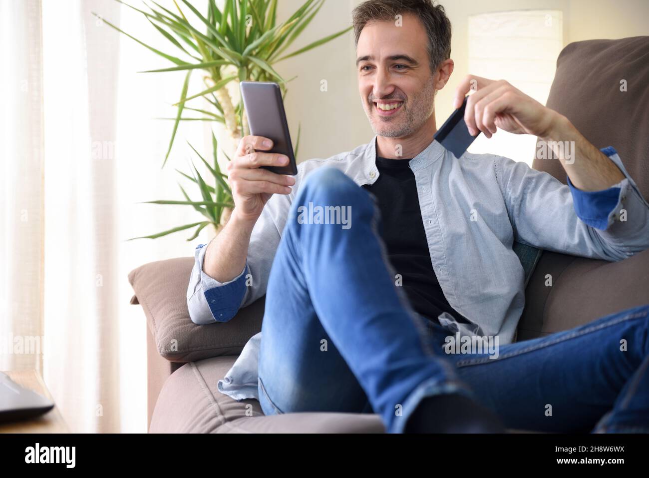 Uomo felice che acquista online da uno smartphone che si trova sul divano a casa. Composizione orizzontale. Foto Stock