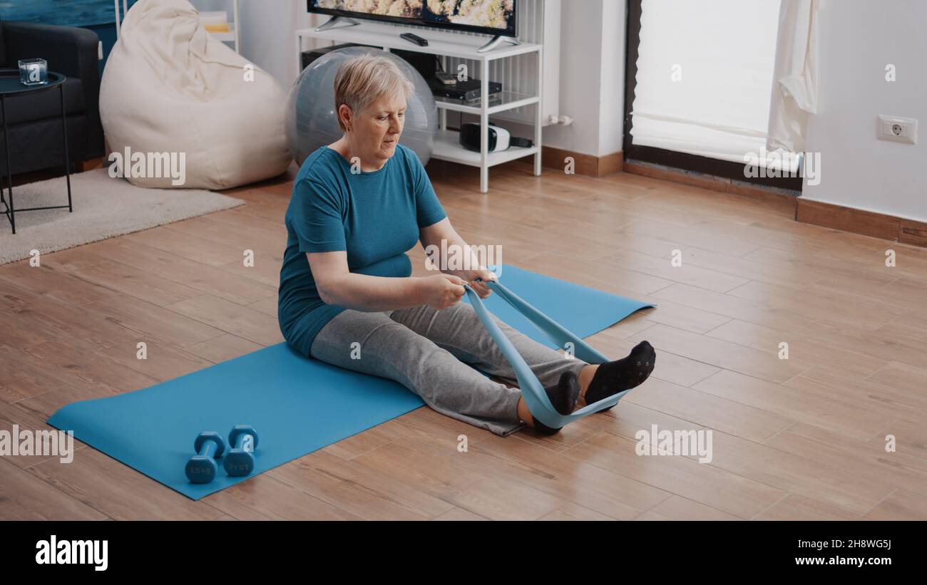 Donna anziana che allunga braccia e gambe con banda di resistenza, seduta  su materassino yoga. Cintura elastica in pensione per adulti per fare  esercizio fisico e sport. Allenamento donna con attrezzature fitness