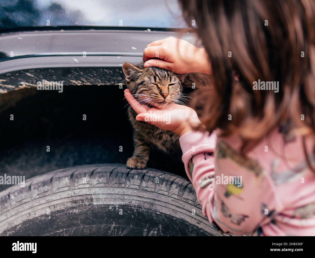 Crop girl in pigiama carezzando adorabile homeless gatto seduto su ruota di pneumatico di auto Foto Stock