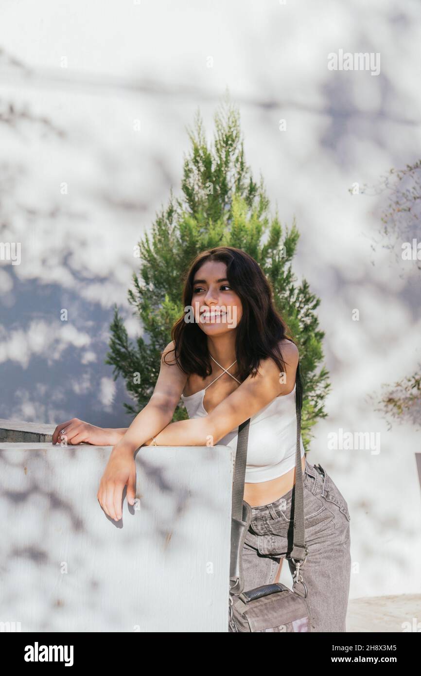 Contenuto Signora ispanica con capelli marroni appoggiati su una barriera di pietra sulla strada contro il muro e guardando via in giornata di sole Foto Stock