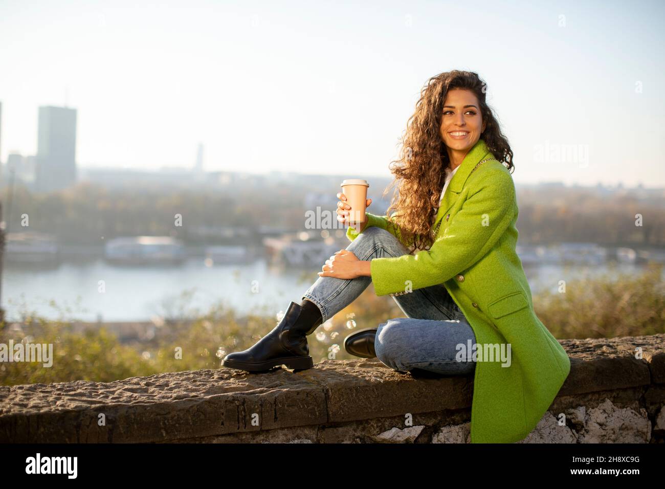 Bella giovane donna capelli ricci godendo il sole autunnale mentre si siede accanto al fiume e bere caffè takeaway Foto Stock