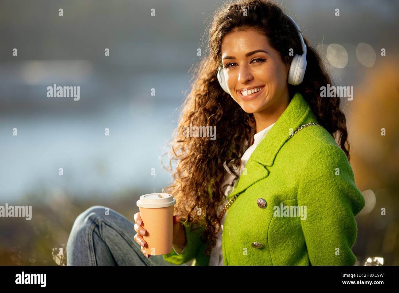 Bella giovane donna che ascolta la musica con smartphone e caffè da asporto sul fiume, nella soleggiata giornata autunnale Foto Stock