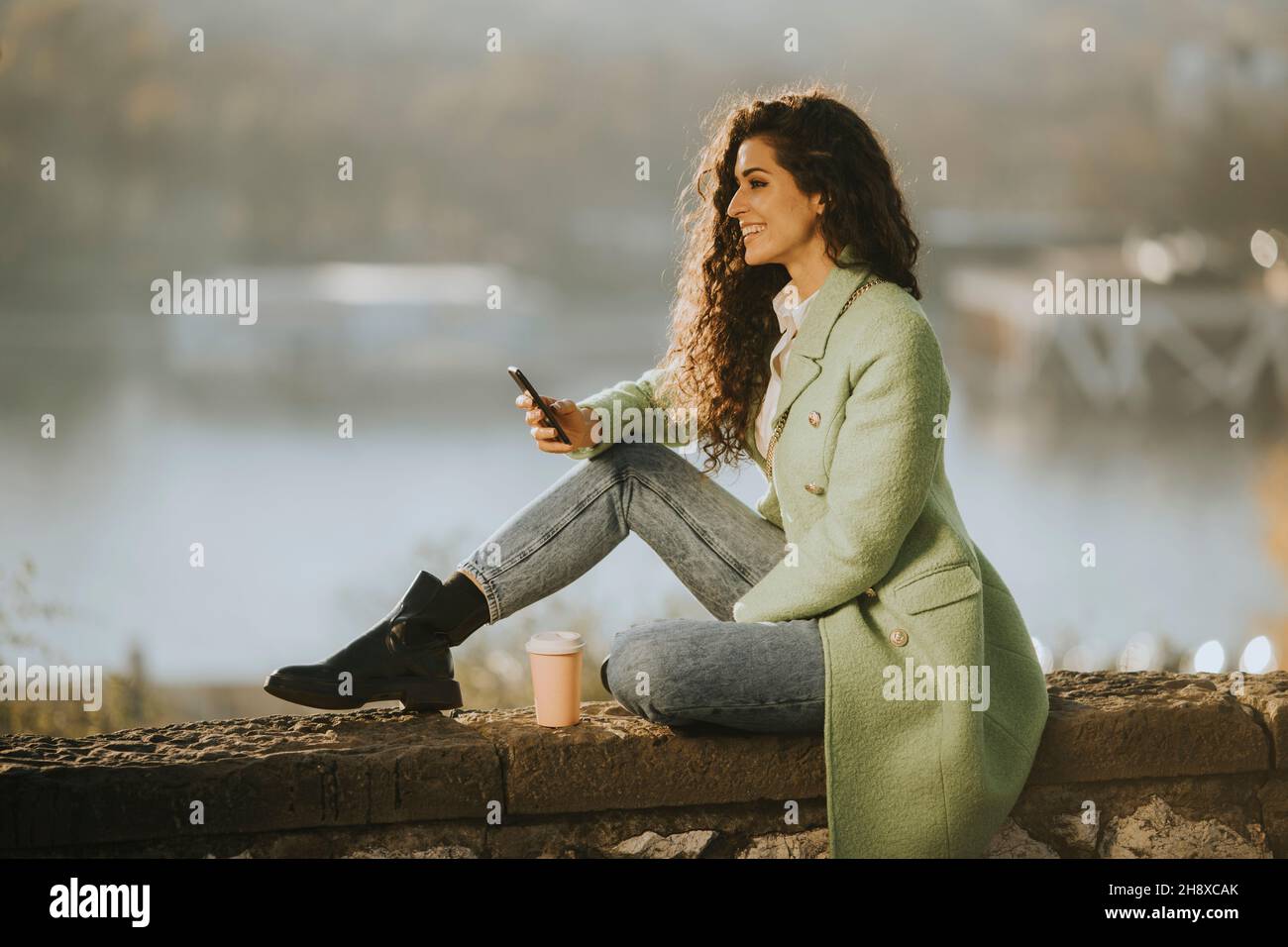 Bella giovane donna ricci capelli usando smartphone mentre si siede accanto al fiume e bere caffè takeaway Foto Stock