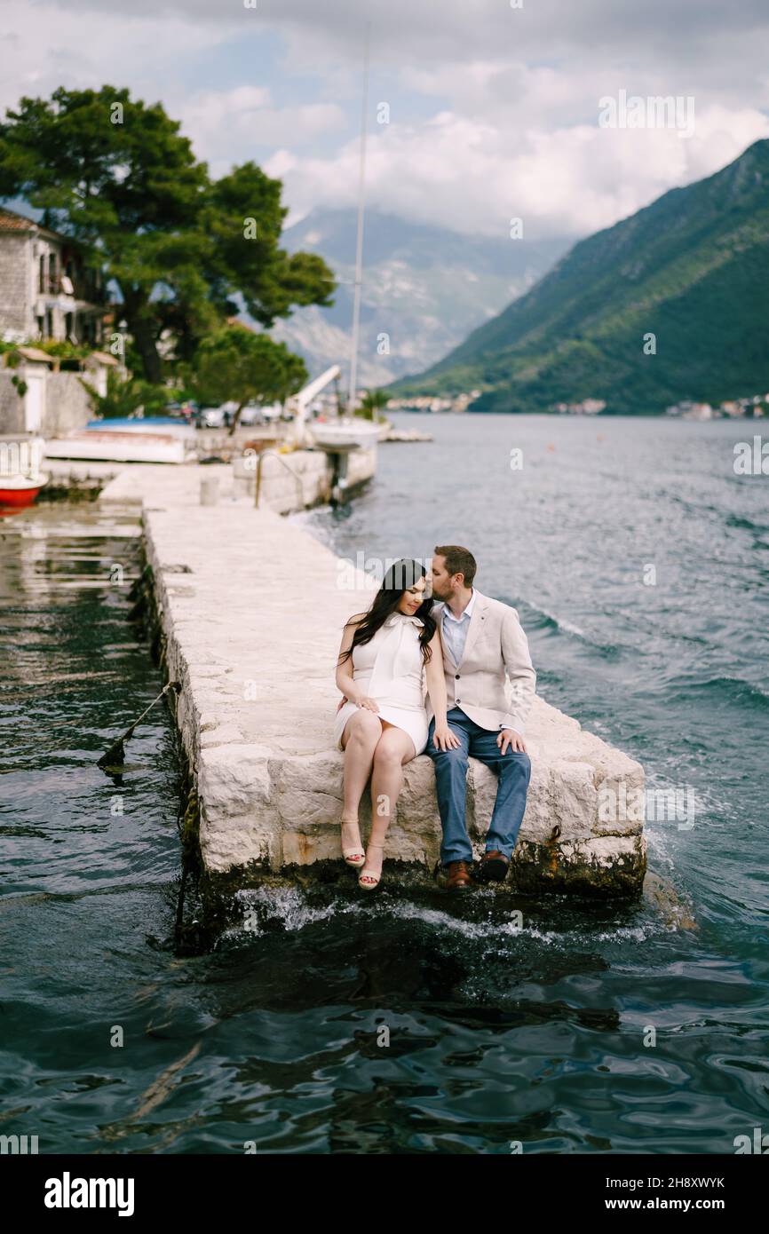 L'uomo bacia la donna sulla fronte mentre si siede sul molo vicino all'acqua Foto Stock