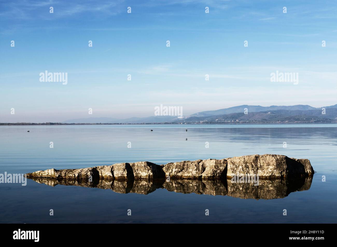 Rocce nelle acque limpide del lago Trasimeno in Italia , sullo sfondo del centro storico Foto Stock