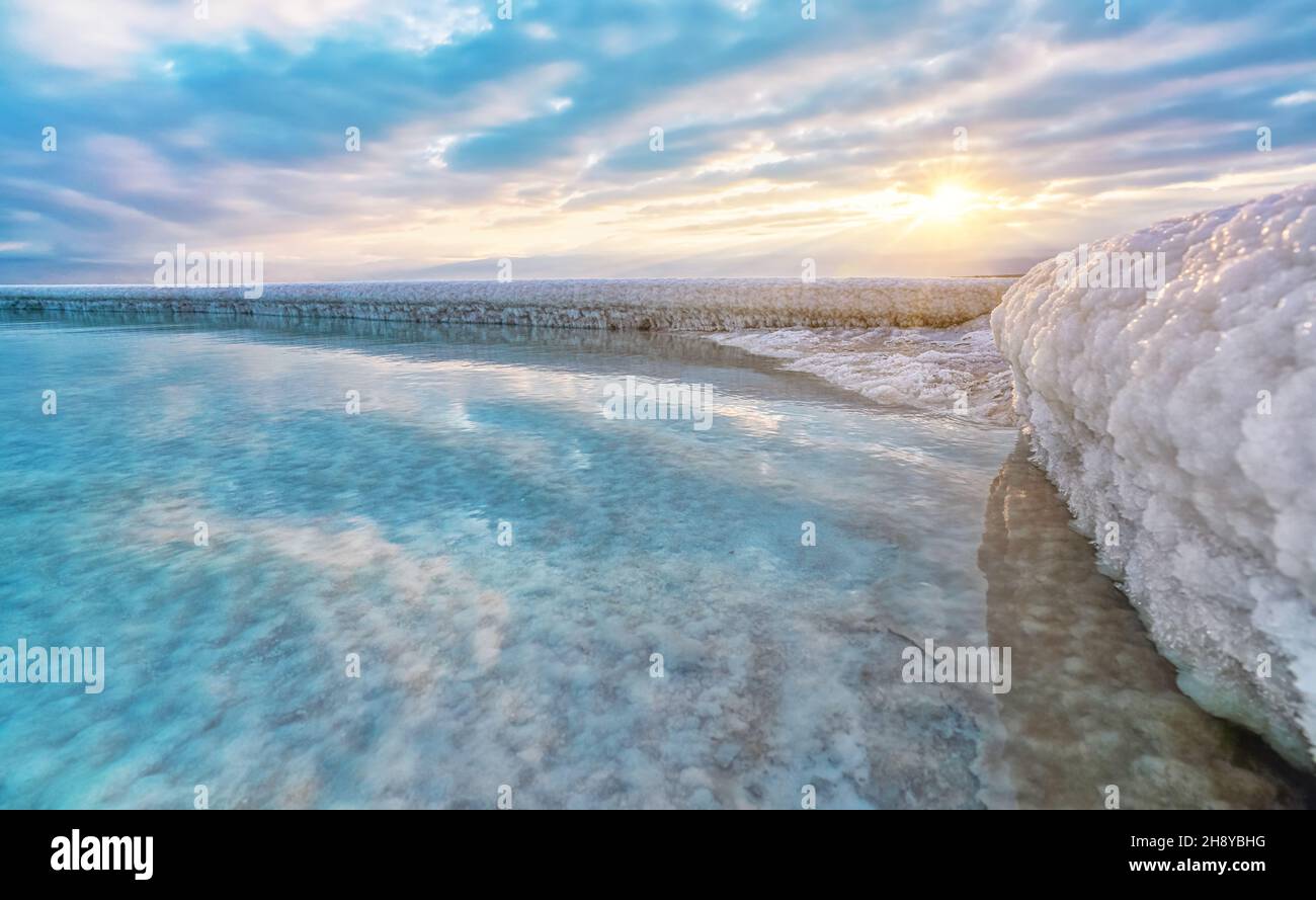 Sabbia completamente ricoperta di sale cristallino assomiglia a ghiaccio o neve sulla riva del Mar Morto, acque turchesi blu vicino, cielo colorato con dis del sole del mattino Foto Stock