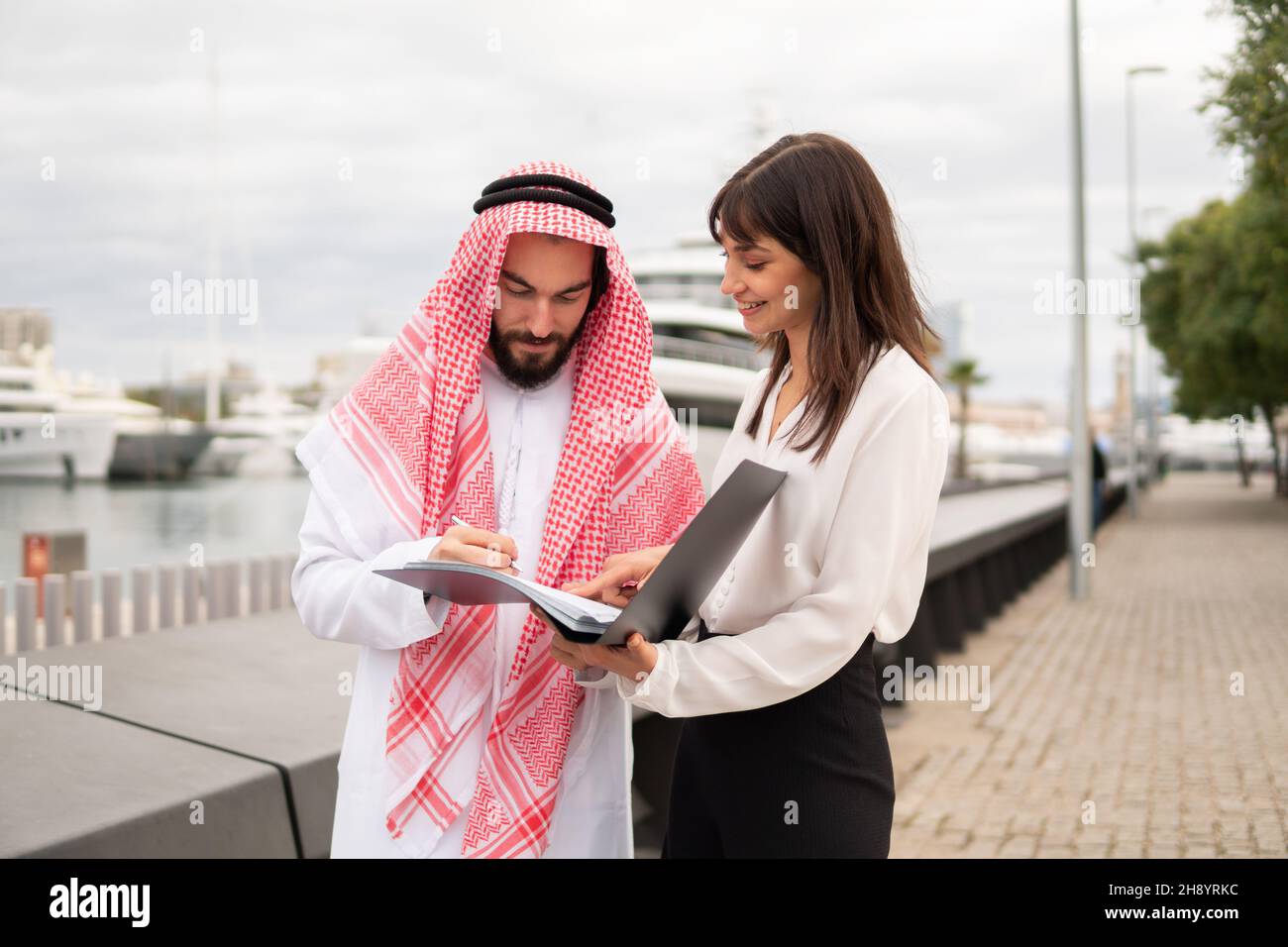Uomo arabo cantando contratto di noleggio per prenotare yacht, uomo d'affari saudita in abbigliamento tradizionale che fa affari con giovane caucasica mediatore mentre si levano in piedi insieme nel porto Foto Stock