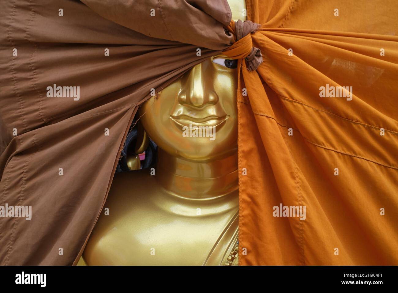 Statua del Buddha in una fabbrica di oggetti buddisti in Bambung Muang Rd., Bangkok, Thailandia, un occhio che si spacca da dietro una copertura di tessuto arancione e marrone Foto Stock