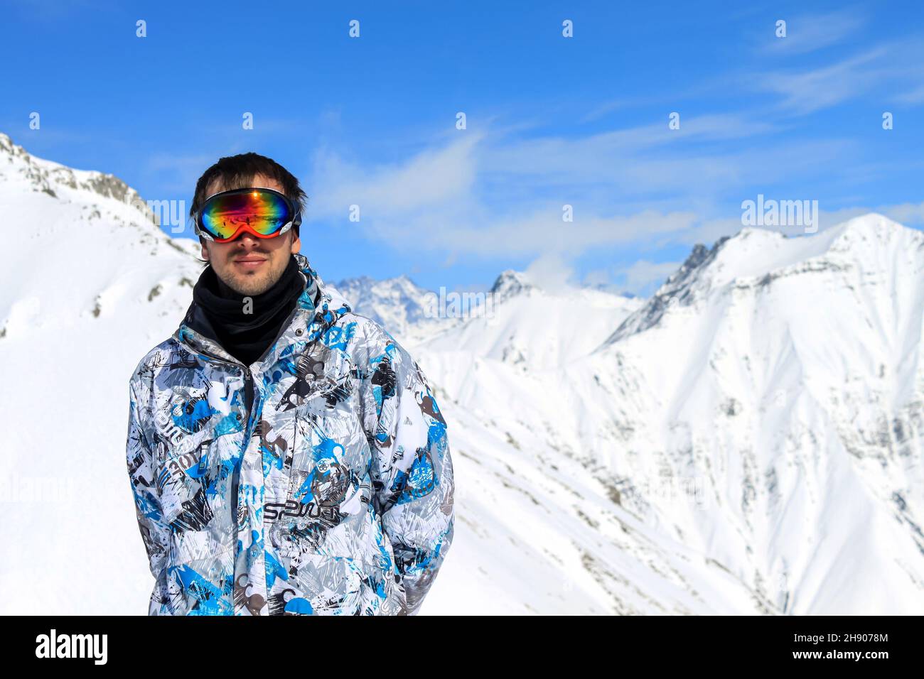 Un ragazzo in una giacca da snowboard e maschera in piedi sullo sfondo di vette innevate di montagna e cielo blu nuvoloso. Vacanze invernali attive sul monte Foto Stock
