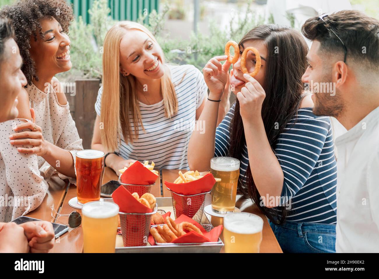 Allegri amici multirazziali in abiti casual divertirsi mentre si siede a tavola con bicchieri di birra e gustosi spuntini sulla terrazza nel caffè di giorno Foto Stock