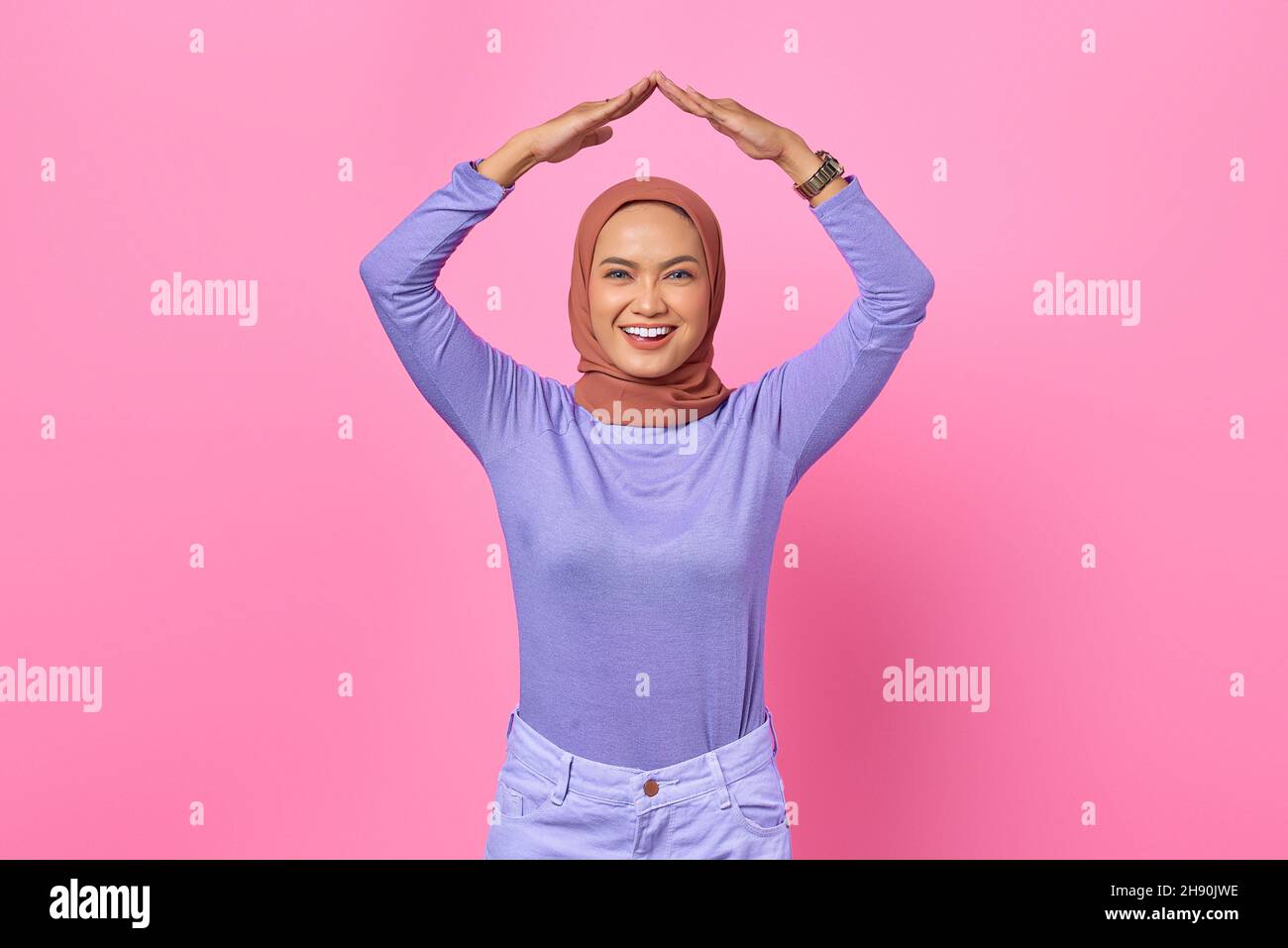 Sorridente giovane donna asiatica che mostra il gesto del tetto con le mani sopra la testa su sfondo rosa Foto Stock