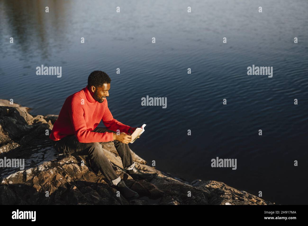 Uomo che legge libro mentre si siede su roccia durante il fine settimana Foto Stock