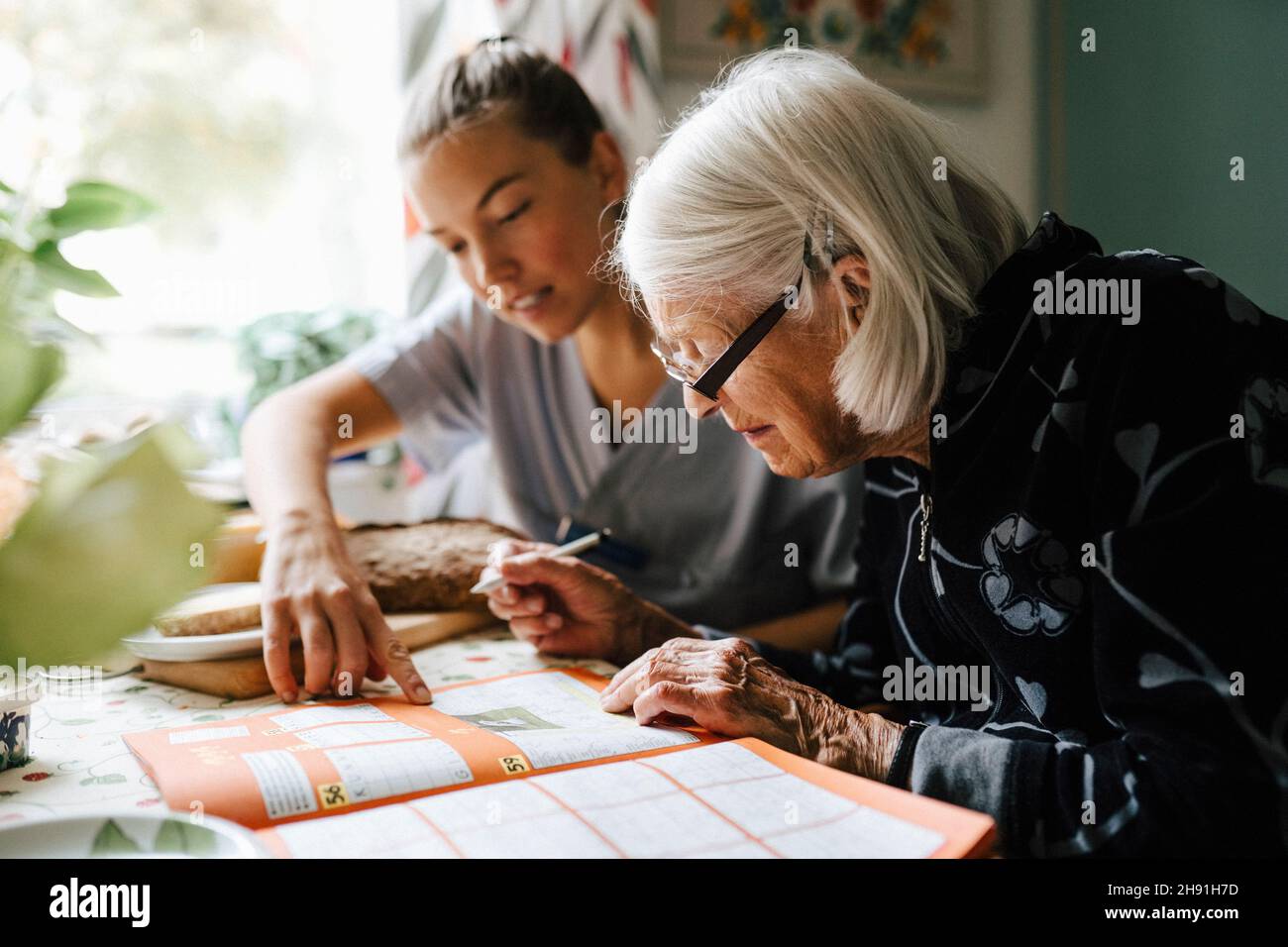 Donna infermiera risolvere puzzle cruciverba con donna anziana mentre si siede in cucina a casa Foto Stock