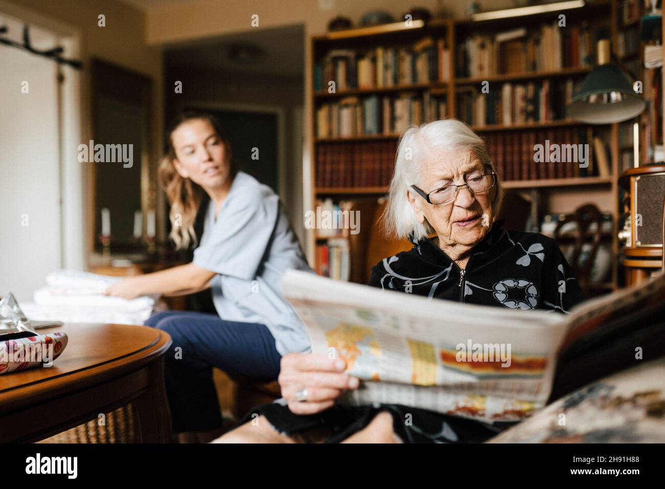 Donna anziana che legge il giornale mentre caregiver femminile che la guarda in salotto Foto Stock