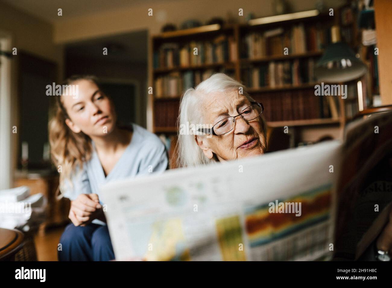 Donna anziana che legge il giornale da parte di un operatore sanitario femminile in soggiorno Foto Stock