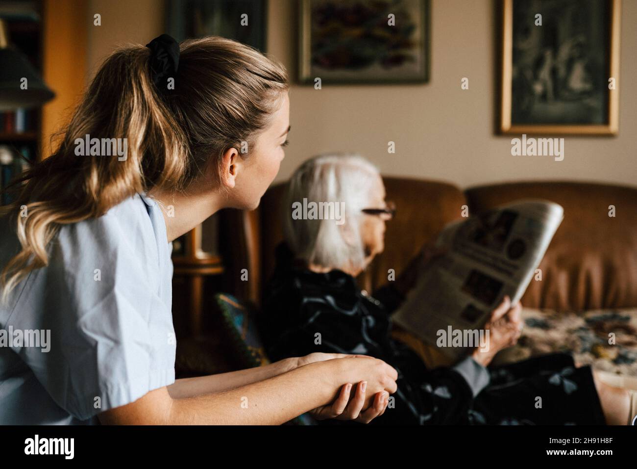Infermiera femminile che guarda la donna anziana che legge il giornale in soggiorno Foto Stock