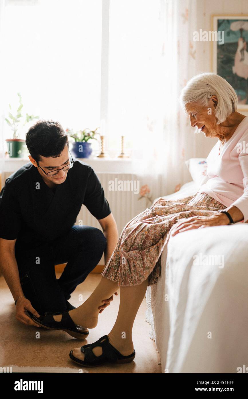 Sorridente donna anziana che guarda mentre l'infermiera maschile la aiuta a  mettere il sandalo in camera da letto Foto stock - Alamy