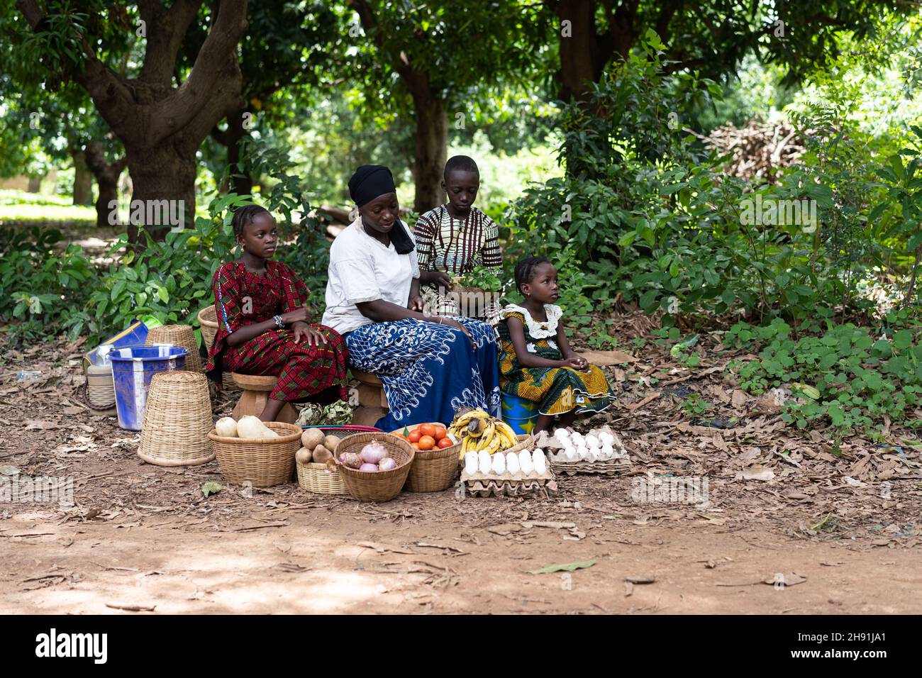 Moglie e figli dell'agricoltore dell'Africa occidentale seduti sul bordo di una strada sterrata in attesa che i clienti acquistino i loro prodotti coltivati in casa Foto Stock