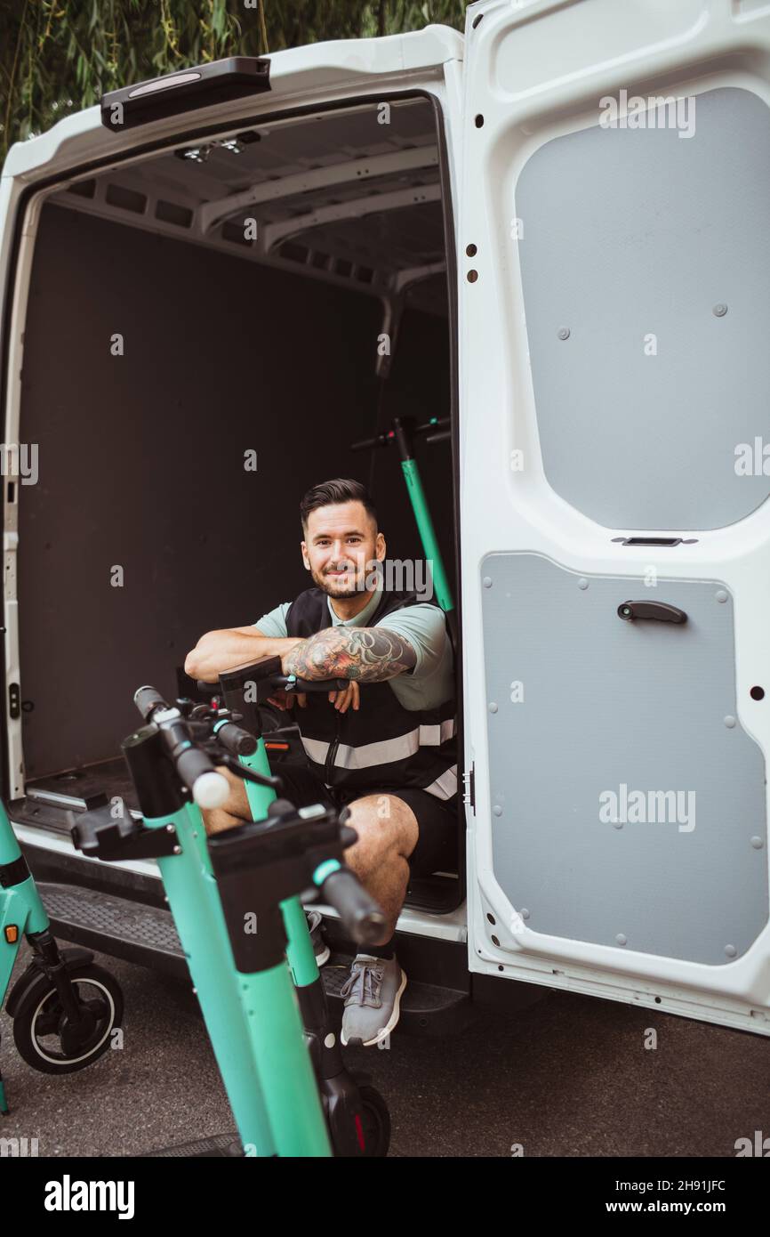 Ritratto di uomo sorridente con colletto blu seduto in furgone di consegna mentre si appoggia su scooter elettrico push Foto Stock