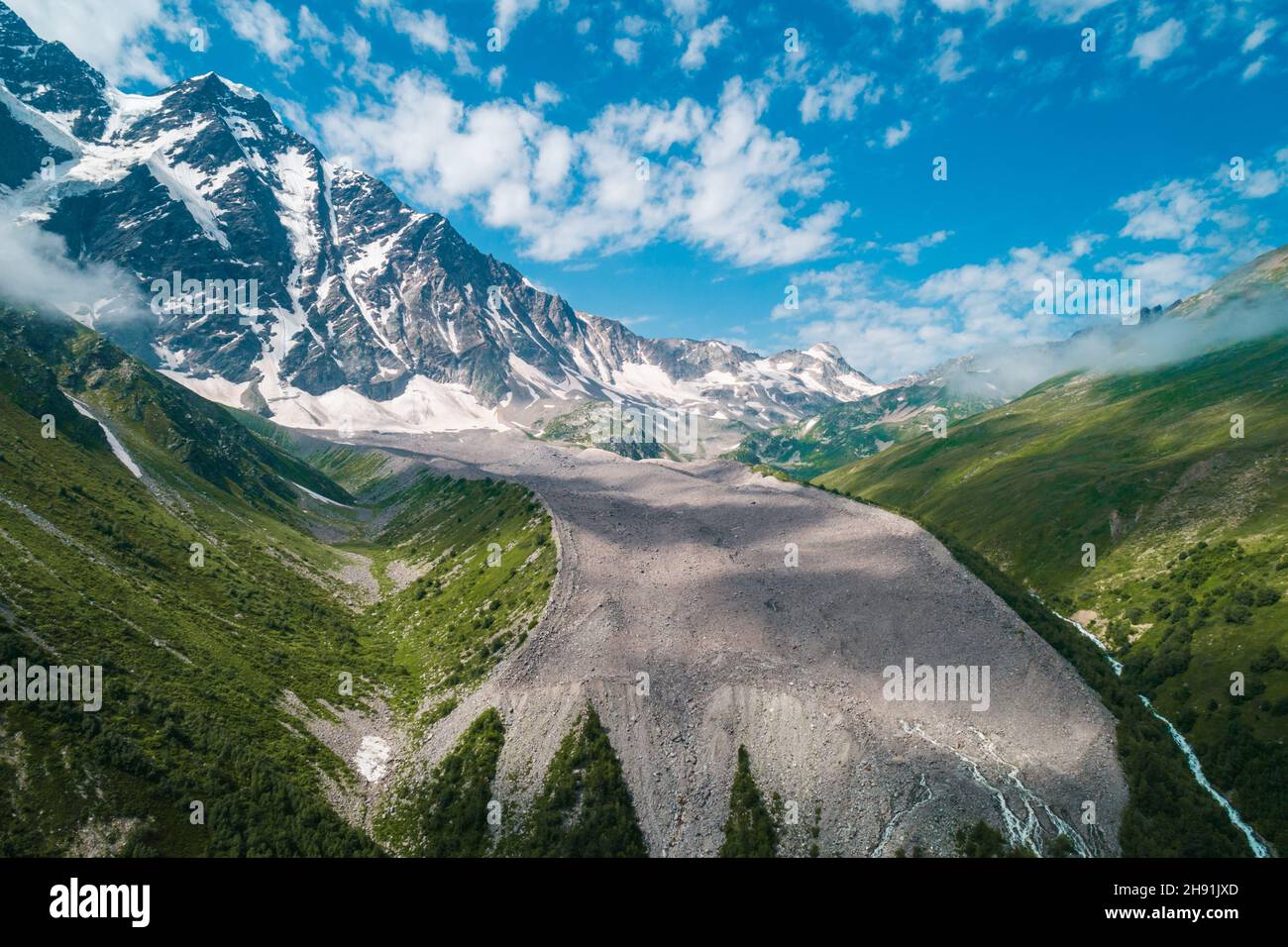 Vette innevate nella giornata estiva soleggiata con il cielo blu e le nuvole sullo sfondo, erba verde di fronte. Bellissimo paesaggio delle montagne del Caucaso. Foto Stock