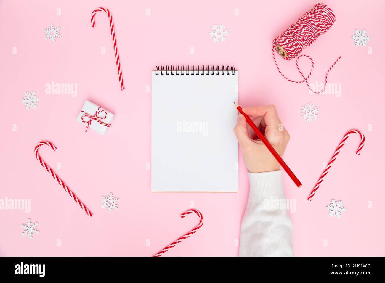 Sfondo accogliente Natale. Scrittura femminile a mano in taccuino bianco con decorazioni festive, scatole regalo su rosa pastello. Pianificazione del nuovo anno, obiettivi, elenco di cose da fare o concetto di lista dei desideri. Disposizione piatta, spazio di copia Foto Stock