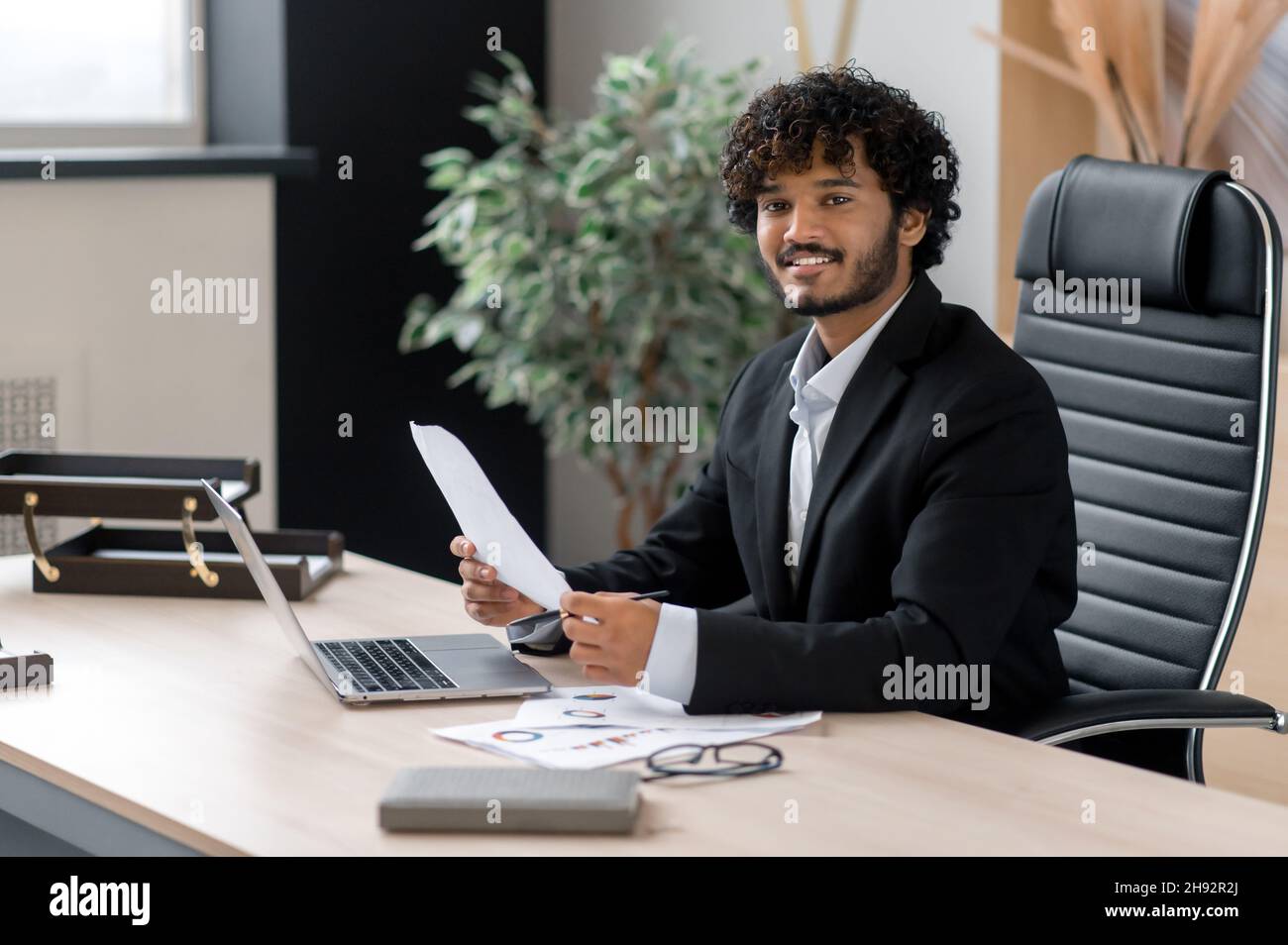 Ritratto di un riuscito uomo d'affari indiano ricci-capelli positivi, vestendo il vestire di affari, seduto alla sua scrivania in ufficio, guardando la macchina fotografica con un sorriso amichevole, tiene grafici di affari Foto Stock