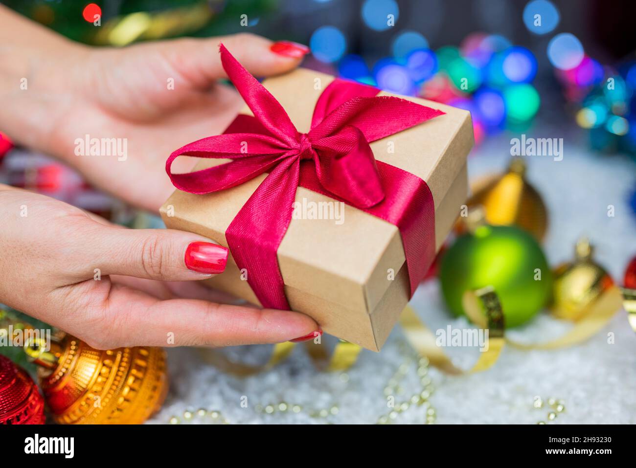 Confezione regalo con un arco nelle mani di una ragazza con una serpentina e giocattoli di Natale sullo sfondo di un albero di Natale e bokeh per il nuovo anno Foto Stock