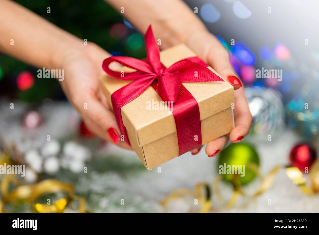 Confezione regalo con un arco nelle mani di una ragazza con una serpentina e giocattoli di Natale sullo sfondo di un albero di Natale e bokeh per il nuovo anno Foto Stock