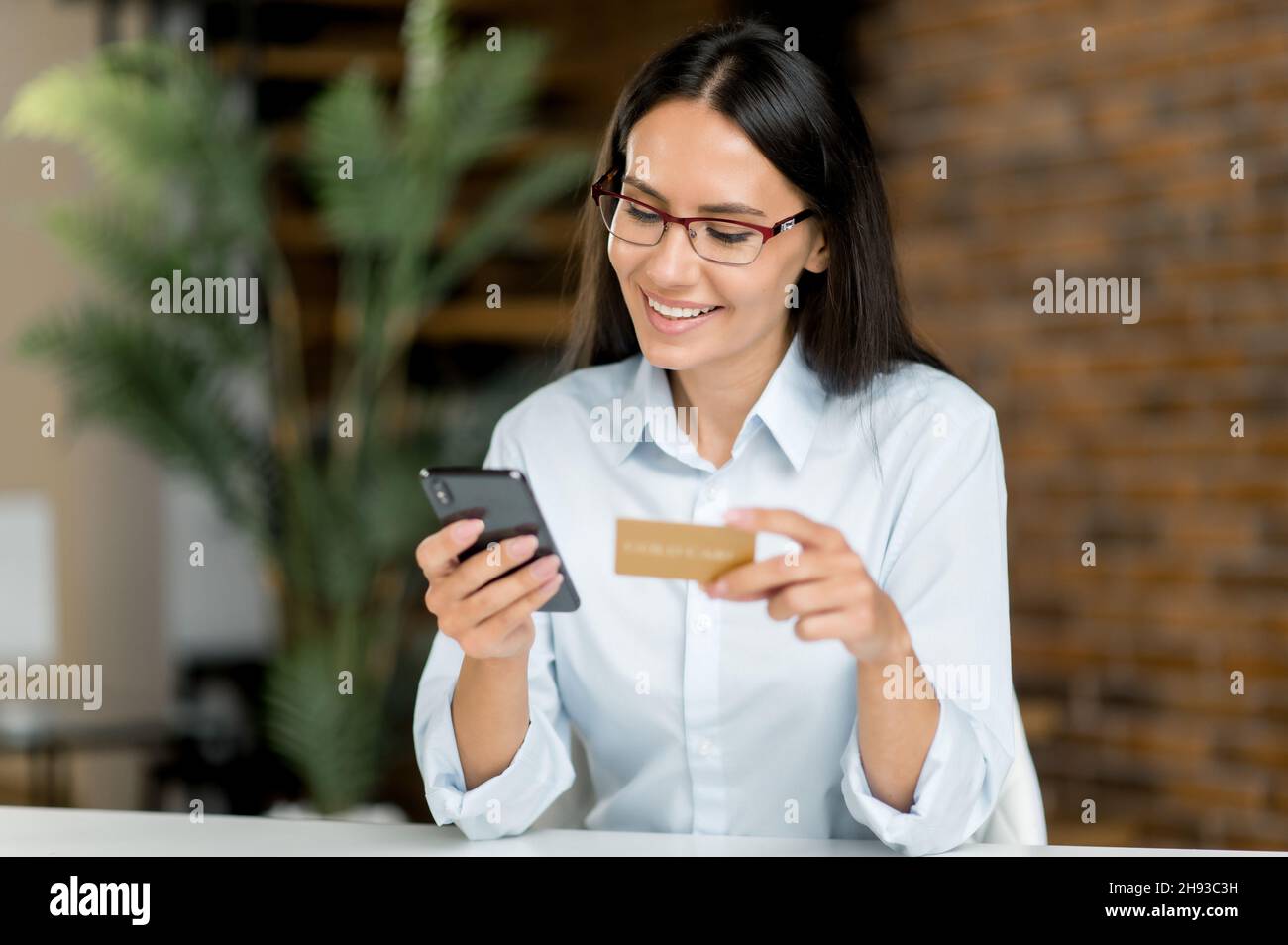 Shopping online. Donna bruna caucasica positiva seduta a un tavolo, usa il cellulare e la carta di credito per pagare gli acquisti online e la consegna, inserisce i dettagli, guarda la carta, sorrisi.transazione online Foto Stock