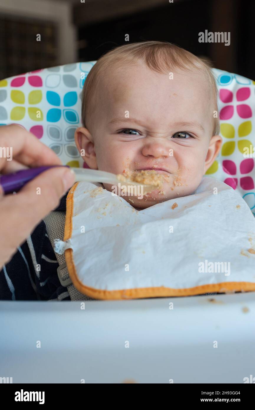 Una bambina (ca. 10 mesi) è stata poppata la sua cena di cibo di purea e rifiuta un cucchiaio con un grimace disgustato. Foto Stock